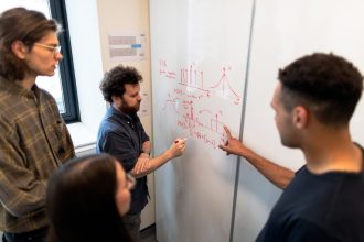 Grupo de engenheiros reúne-se diante de um quadro branco para discutir dados em um ambiente de pesquisa.