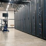 Sala de data center com gabinetes metálicos, servidores, equipamentos de rede e cabos organizados.